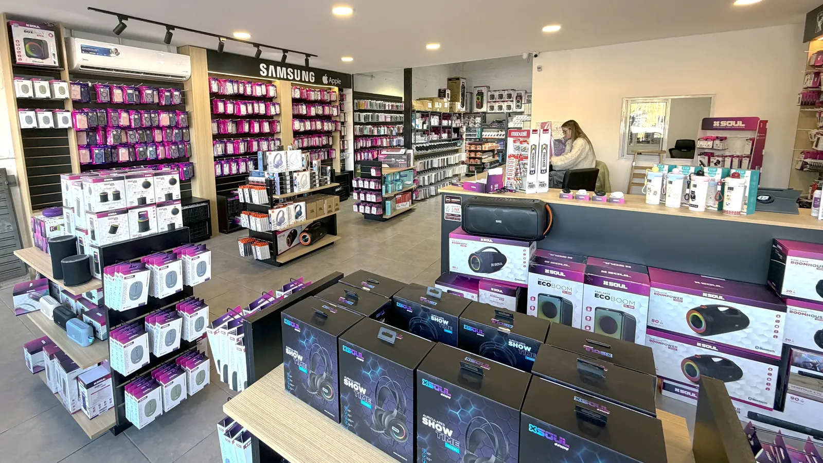 Góndolas del salón mayorista cargadas de parlantes, auriculares y fundas, con el sector Samsung y Apple al fondo